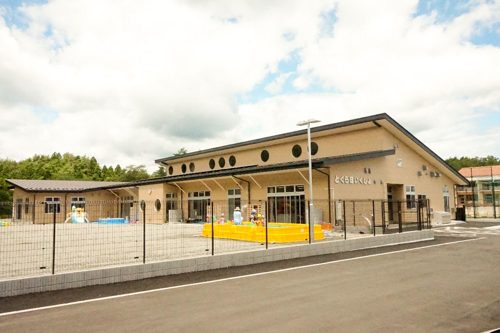 ベージュ色の外壁と黒い屋根で、屋根付近に複数の丸い窓が並んだ建物の前に、フェンスで囲まれた園庭がある戸倉保育所の写真