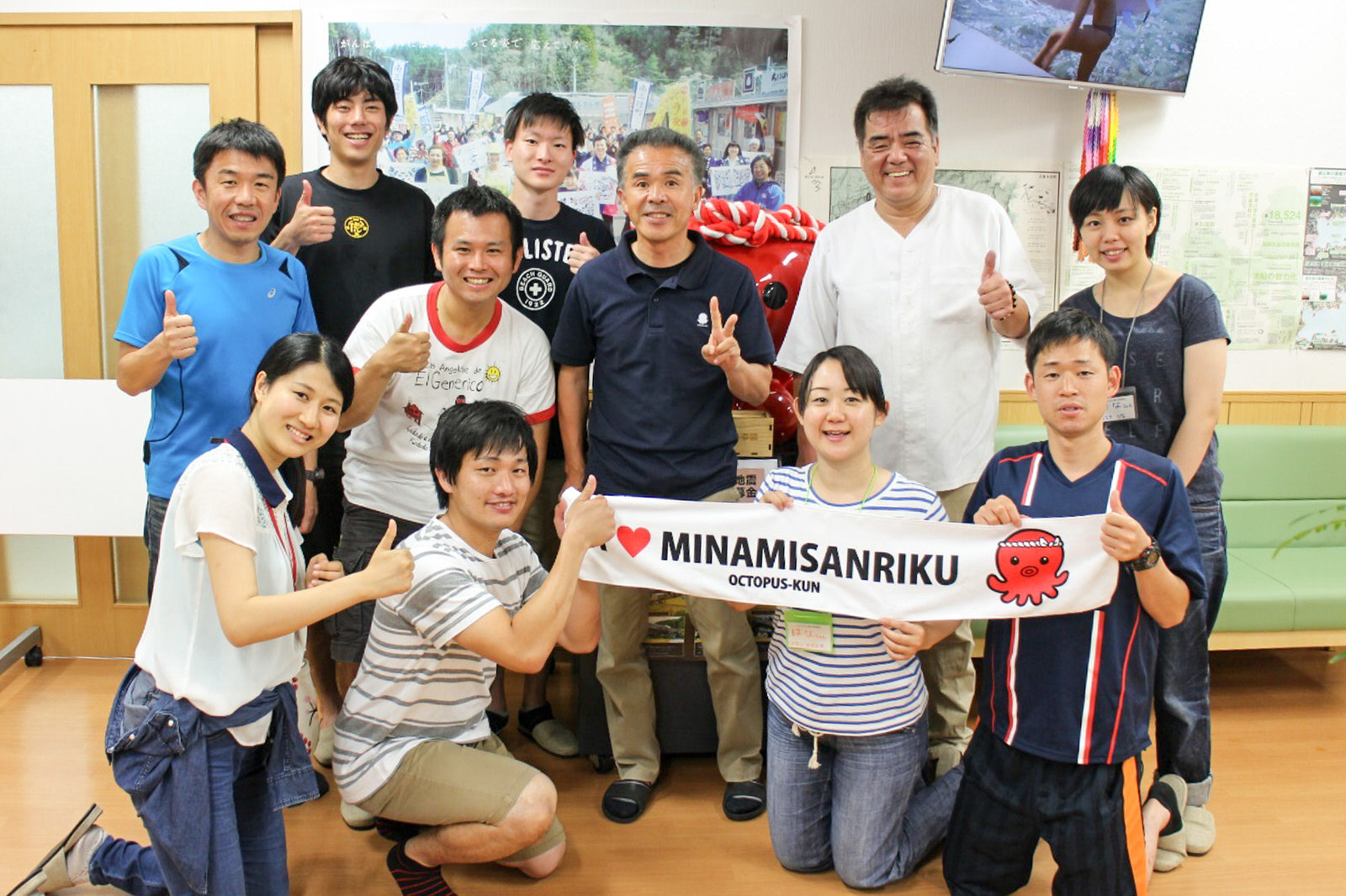 大きく引き伸ばされた写真が壁に飾られた屋内のスペースで、オクトパス君のイラストと「MINAMISANRIKU」と書かれた細い横断幕を手に、メンバーや地元の方が一緒に並んでポーズをとっている写真
