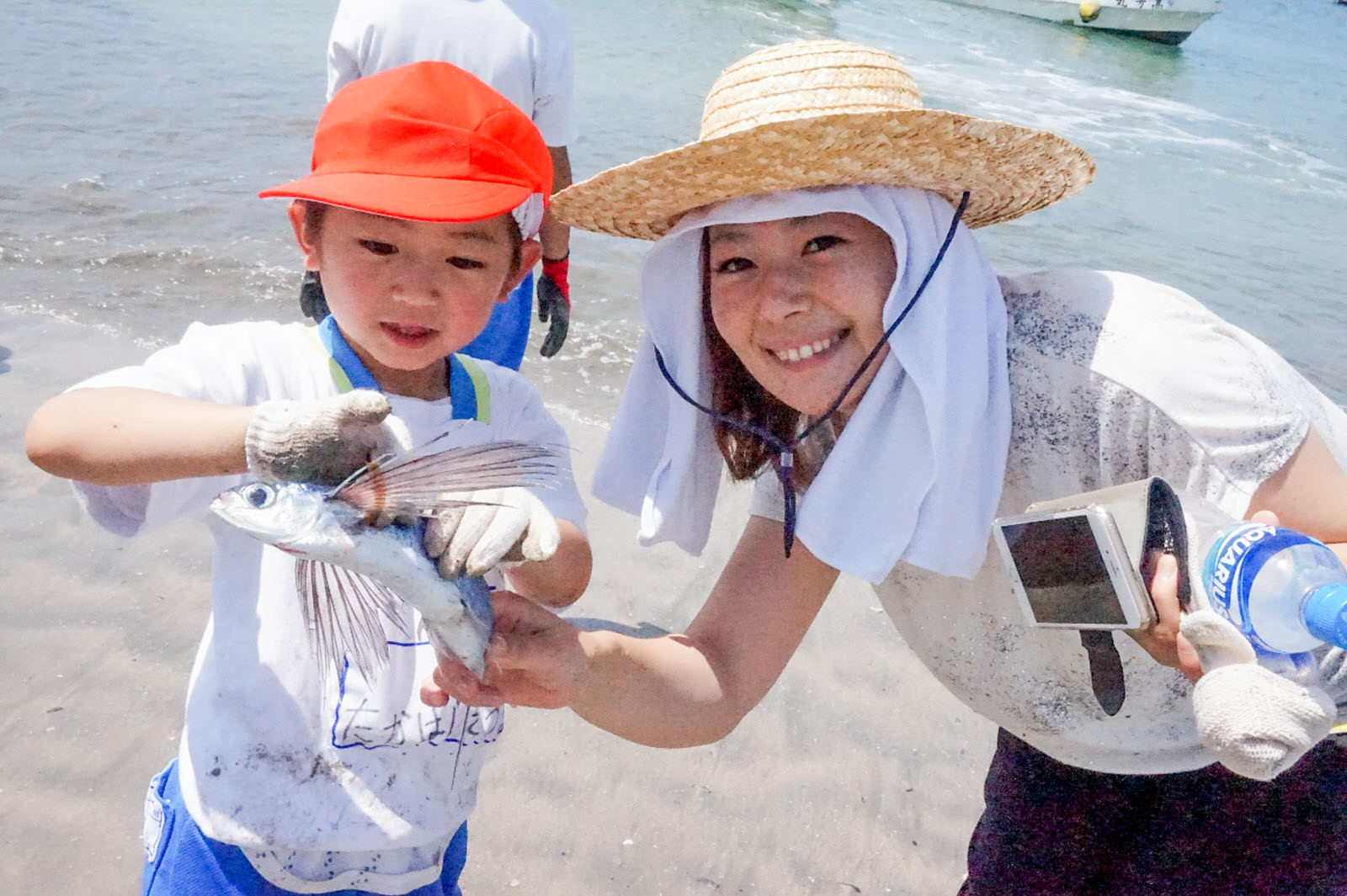 赤い帽子をかぶった男の子が長いヒレをもつ魚を両手で持ち、麦わら帽子に白いタオルを巻いた女性が横で笑顔を向けている浜辺での記念写真
