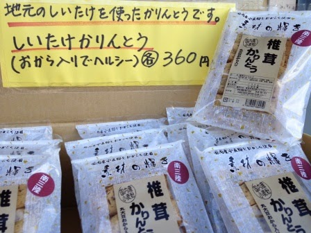 上部に手書きで「地元のしいたけを使ったかりんとうです。」と書かれた黄色いポップが貼られている段ボールの中に、「椎茸かりんとう」と書かれた商品が透明な袋に個包装されて多数並んでいる写真