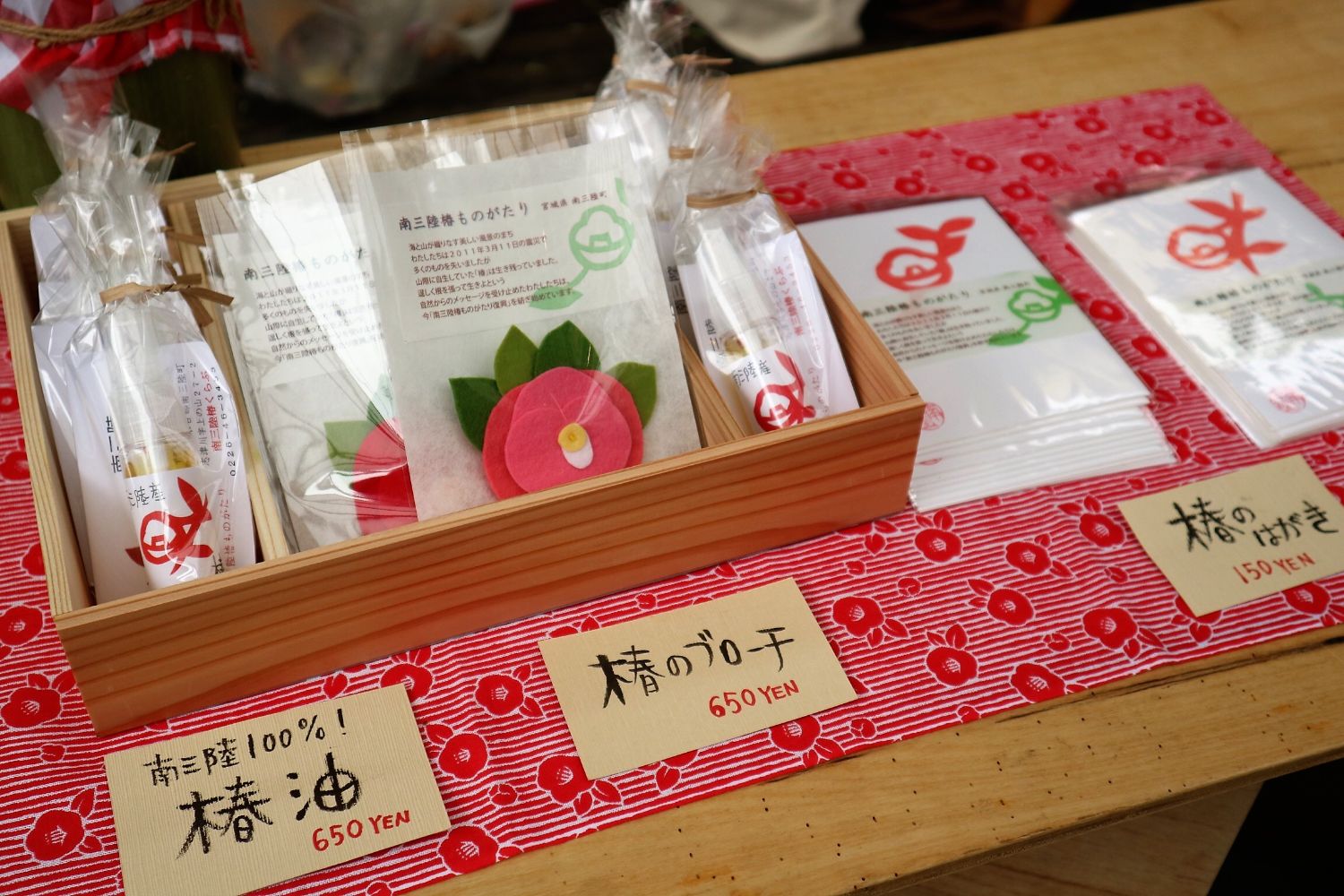 木箱の中に包装された椿製品が整然と並べられ、それぞれに「椿油」「椿のブローチ」「椿のはがき」などと手書きで書かれた値札がついている特産品展示の写真