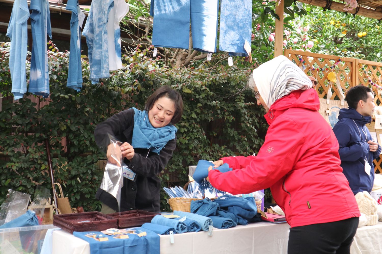 藍染めされた青いストールを巻いた女性スタッフが、藍染めの布を販売しながら笑顔で接客し、買い物客の女性が商品を手に取っている藍染製品のブースの写真