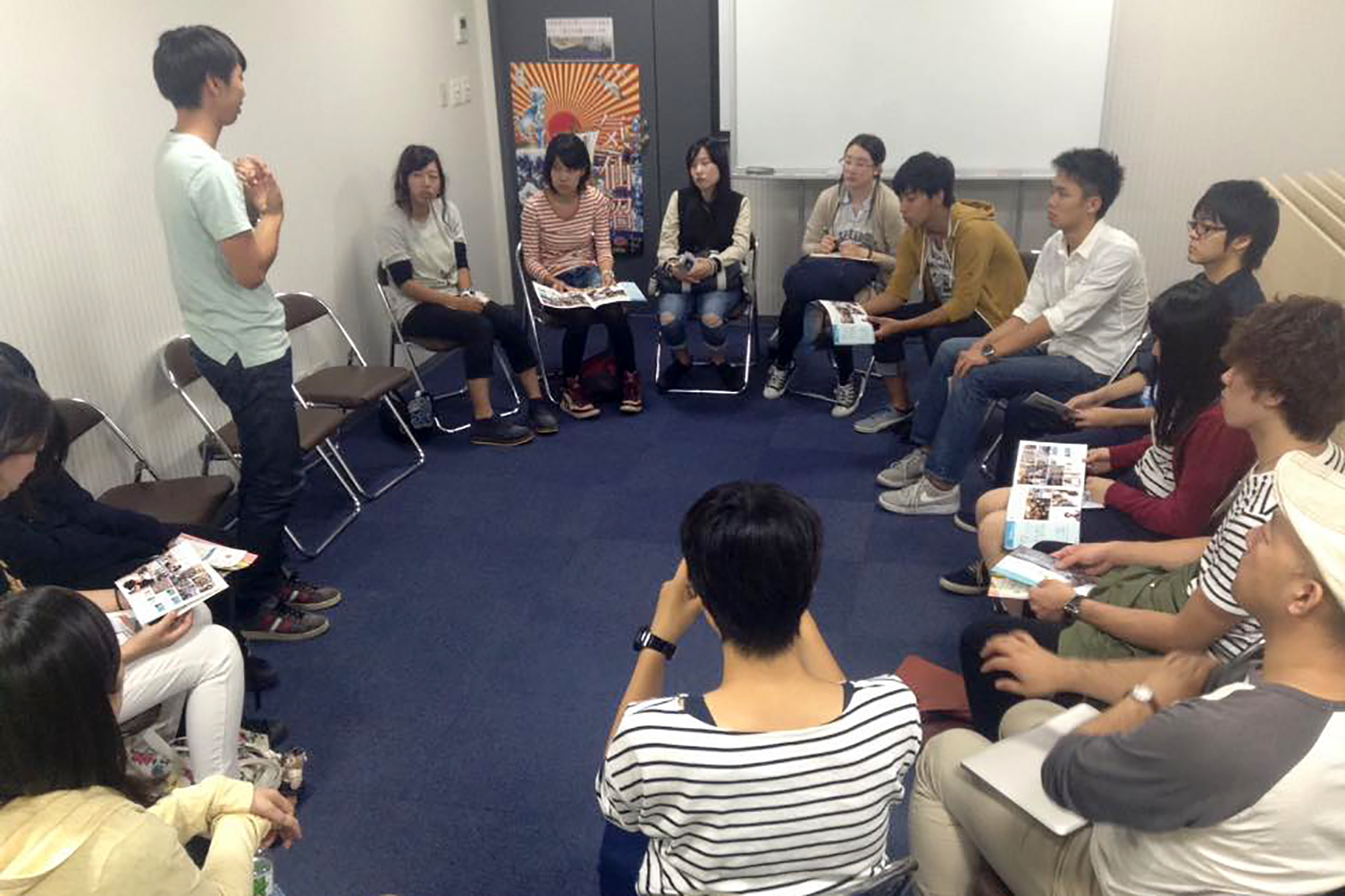 室内で十数人の若者たちが椅子を円形に並べて座り、立って話す若い男性に注目しながらパンフレットを手にしている写真