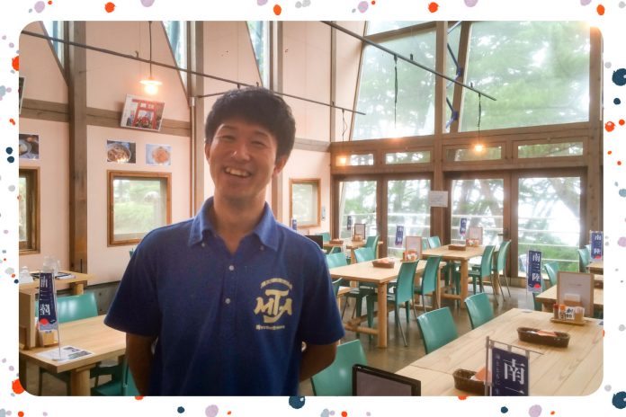 青いポロシャツを着た笑顔の男性が、木のテーブルと緑色の椅子が並ぶガラス張りの明るいレストランの中央に立っている写真にカラフルな縁取りがされている画像