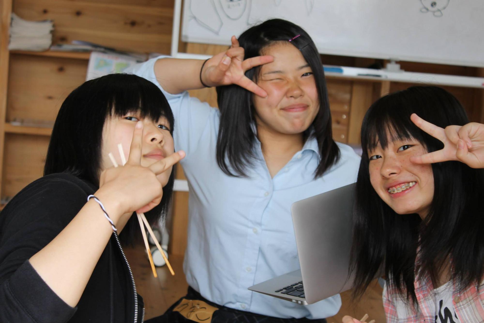 ホワイトボードがある木の部屋の中で、女子高生らしき3人がカメラに向かってピースをしている写真