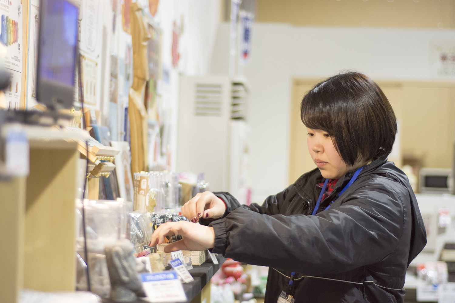 室内の事務所で、観光協会ジャンパーを着た女性が商品棚に手を伸ばして作業している写真