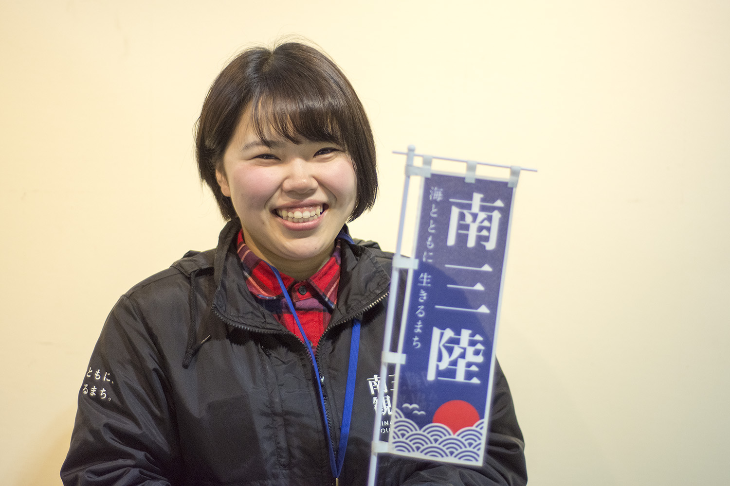 黒の観光協会のジャンパーを着た女性が「南三陸」と書かれたミニのぼり旗を持って笑顔を見せている写真