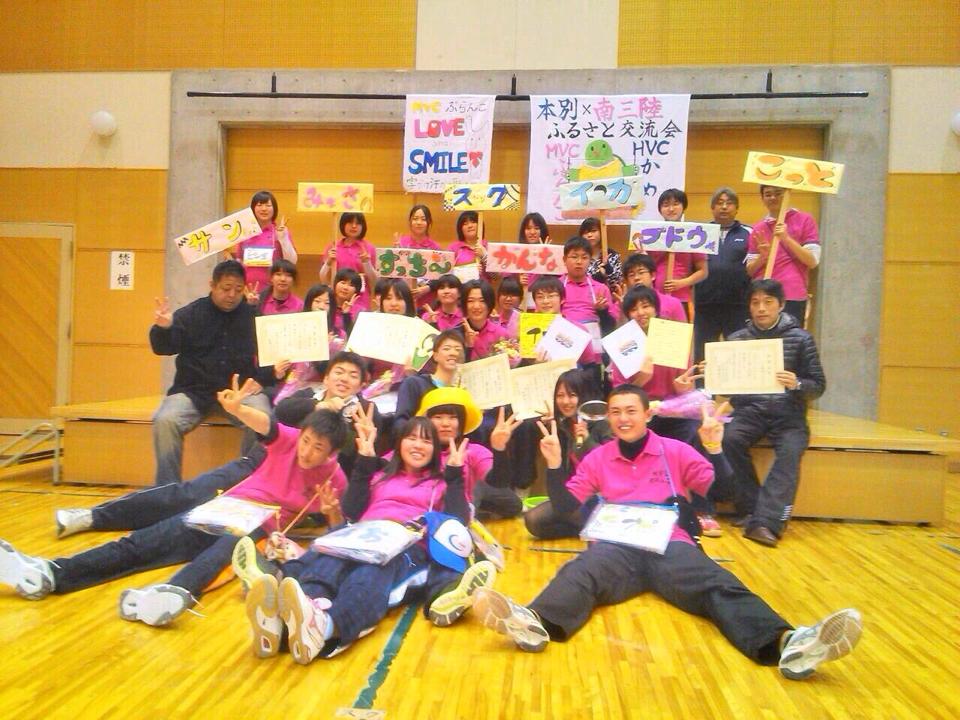 体育館のステージ前で、お揃いのピンクのTシャツを着た大勢の人達がプラカードを掲げて集合写真を撮っている写真