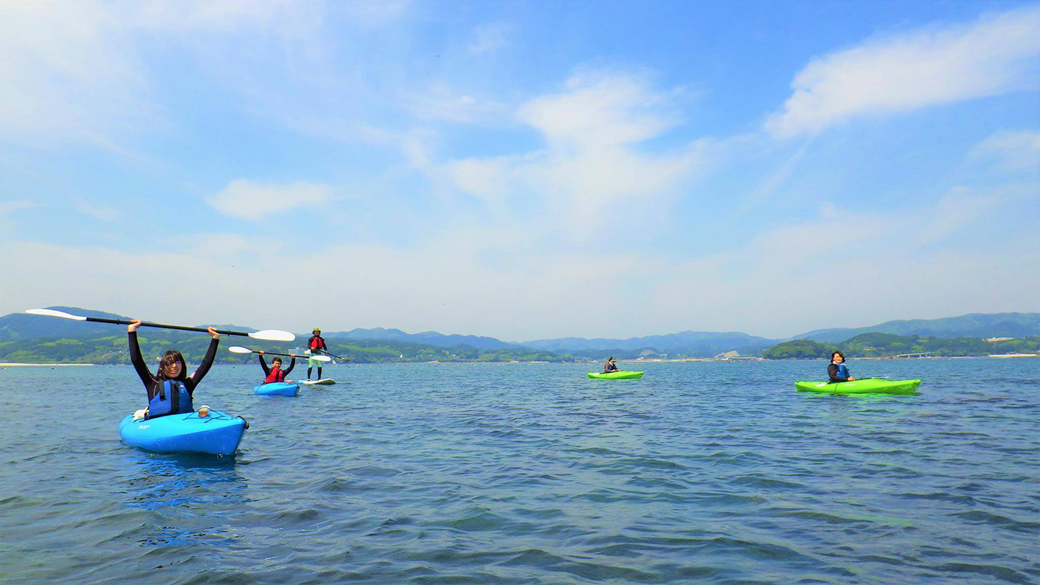 山と青空が見える海の上で、手を挙げてパドルを持ち上げている人もいる青や緑のカヤックに乗った数人の人物が穏やかな海の上に広がって浮かんでいる写真