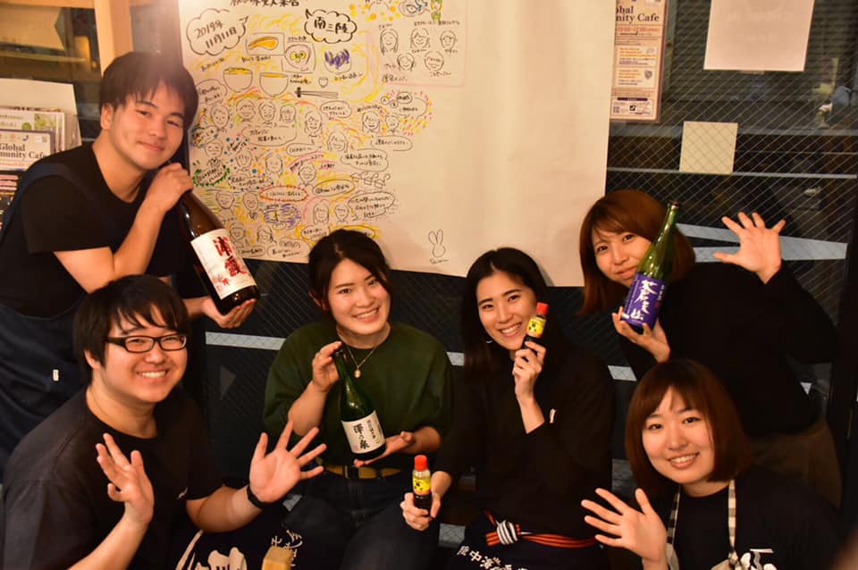 4人が日本酒の瓶や調味料のボトルを手にしており、カメラに向かって手を振ったり、ポーズをとっている「きっかけ食堂」の運営メンバー6人の写真