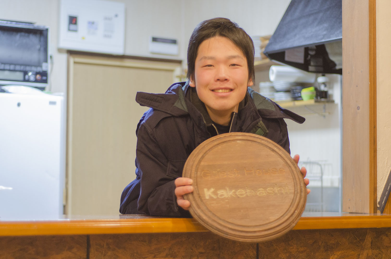 「Guest House Kakehashi」と書かれた木製の丸い看板を持ち、笑顔でカウンター越しに見せる小出悟さんの写真