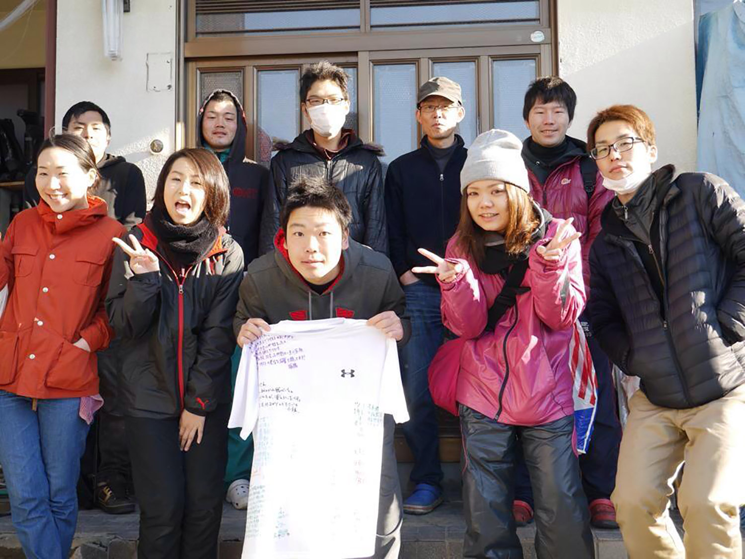 シェアハウスの前で、小出さんがメッセージ入りの白いTシャツを持ち、複数の若者たちが集まって笑顔で写っている集合写真