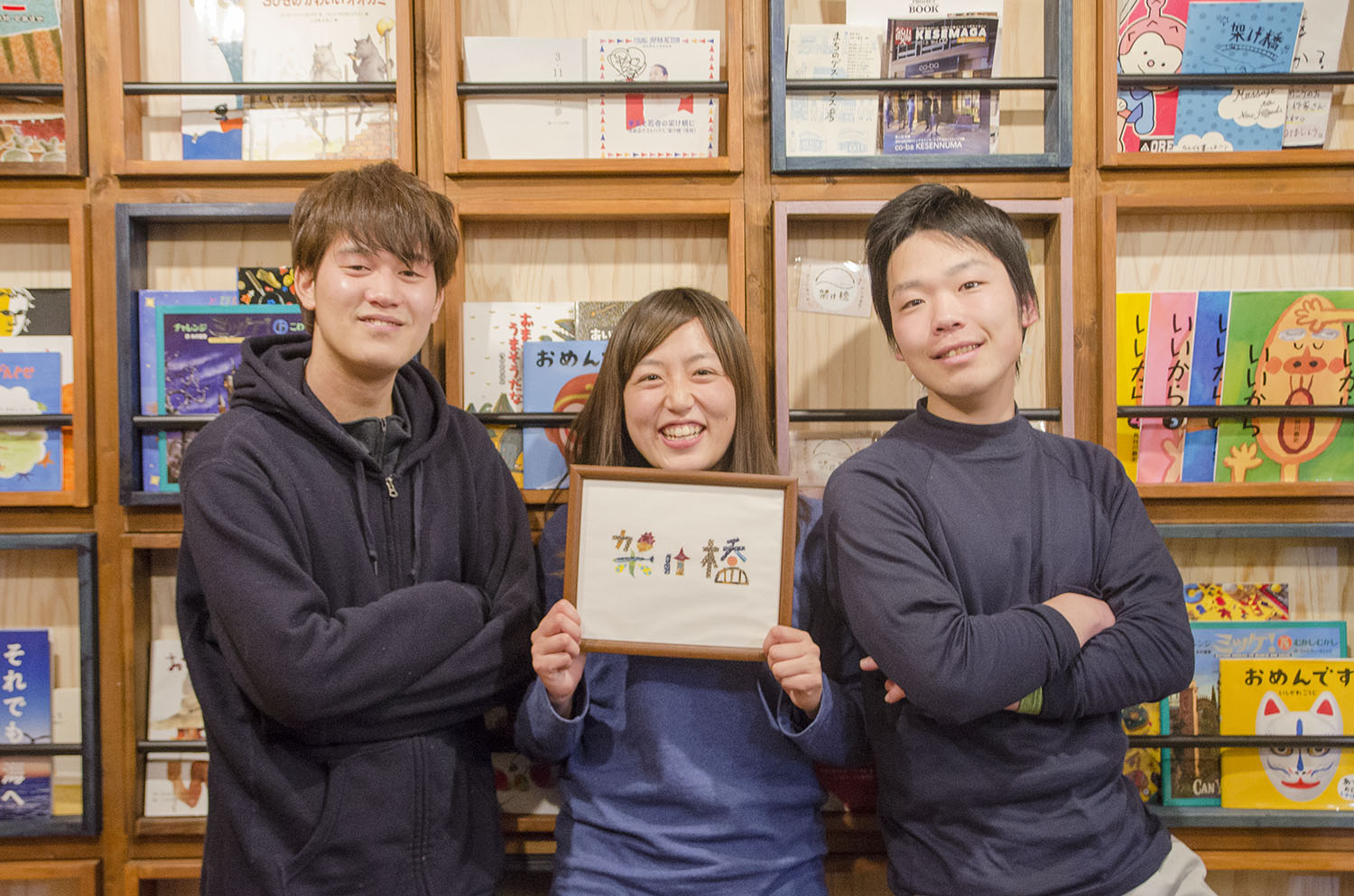 小出さんと2人のスタッフが本棚の前で笑顔で立ち、中央の女性がカラフルな『架け橋』の文字の額縁を持っている写真