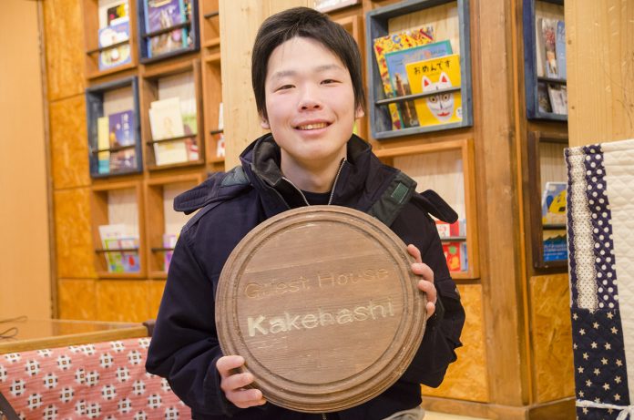 「Guest House Kakehashi」と書かれている木製の丸い看板をもって笑顔で写っている小出悟さんの写真