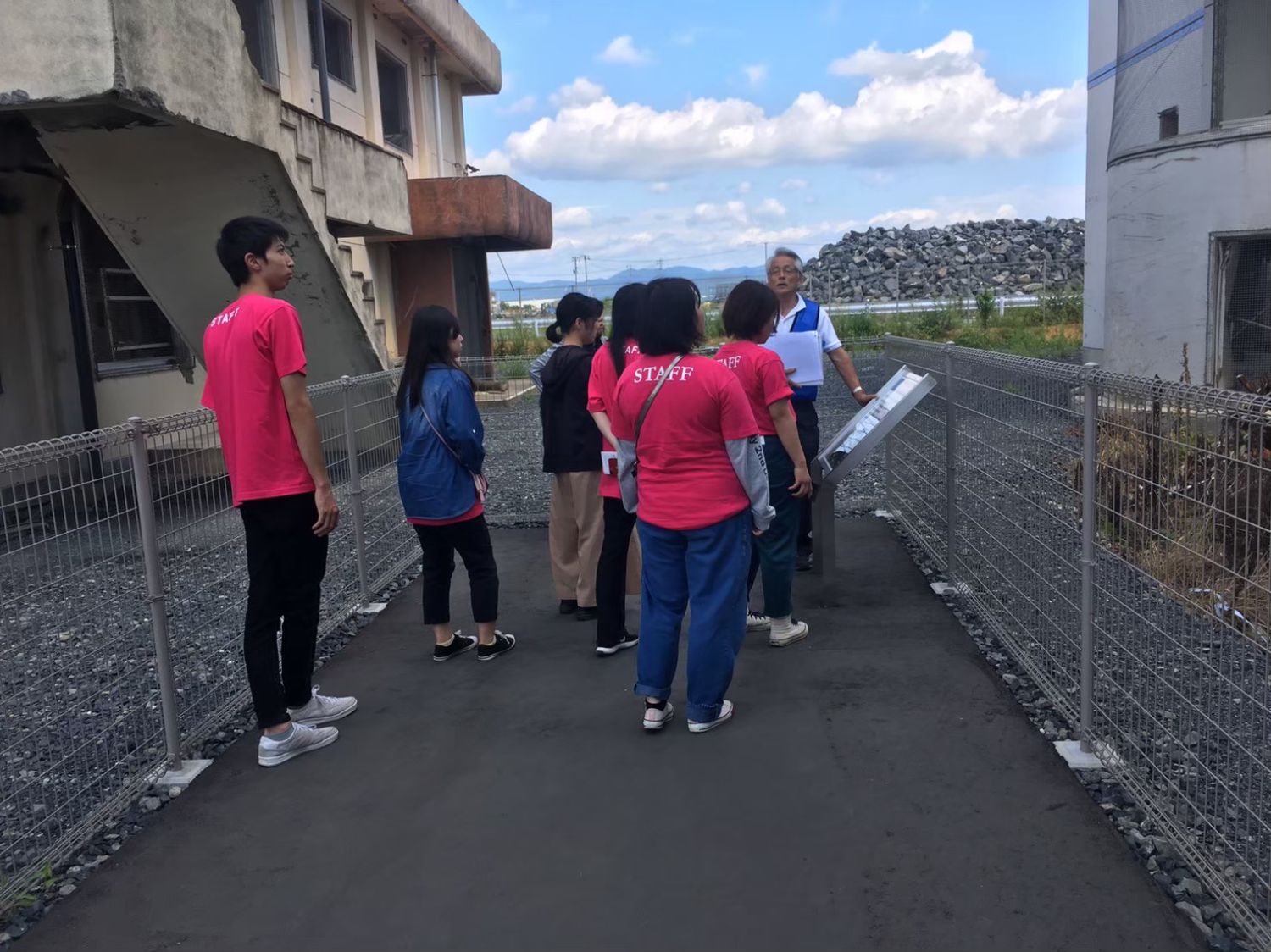 震災遺構前で案内板を見ながら男性の説明を聞く大学生たちの写真