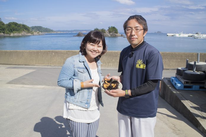 リポーターの女性と高橋さんが海辺で殻付きのまま割ったウニが乗った皿を手に持って笑顔でカメラに見せている写真