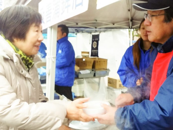 女性がイベント会場のブースで男性スタッフから湯気の立つ温かい食べ物を両手で受け取り笑顔で会話している様子の写真
