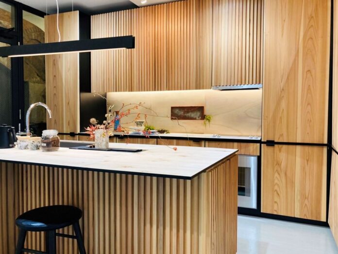 木目調の板で統一された温かみのあるキッチンに、明るい色合いの天板を備えたアイランドカウンターと、その前に置かれた黒い丸椅子がある写真