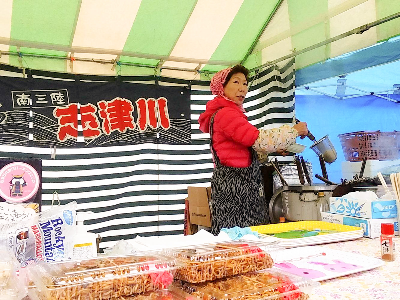 焼きそばのパックが並んでいる飲食の販売ブースで、赤い三角巾に上着を着た女性が調理器具を手にし麺を湯がいている様子の写真
