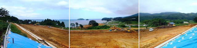青いシートで覆われた法面があり、重機が何台も動き回る造成地が広がり、奥には海と島が見えるパノラマ写真