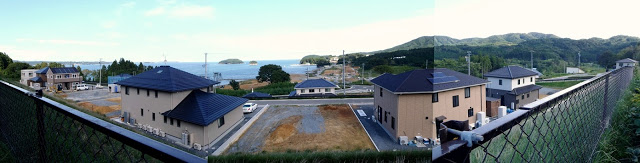 造成された高台の上に新築の住宅が建ち並び、道路も整備されており、奥には海と小島が見えるパノラマ写真