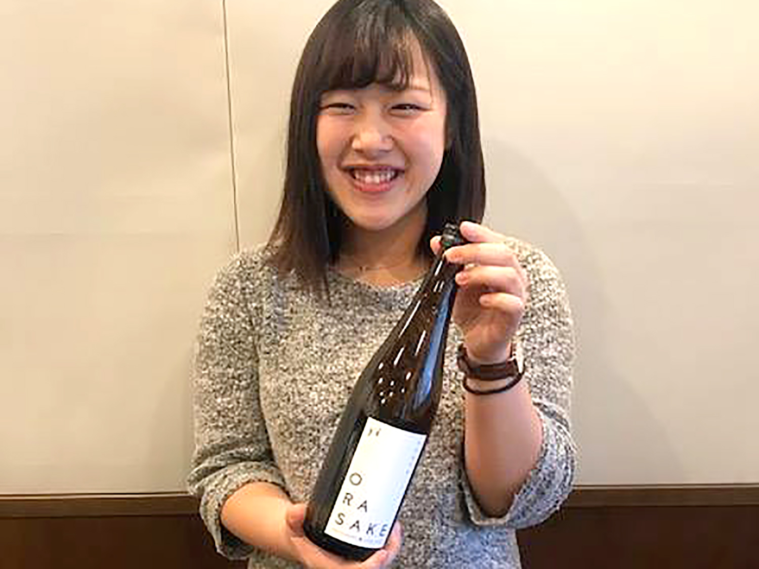 「ORA SAKE」と書かれたラベルの日本酒の瓶を両手で持った佐藤さんが笑顔を見せている写真