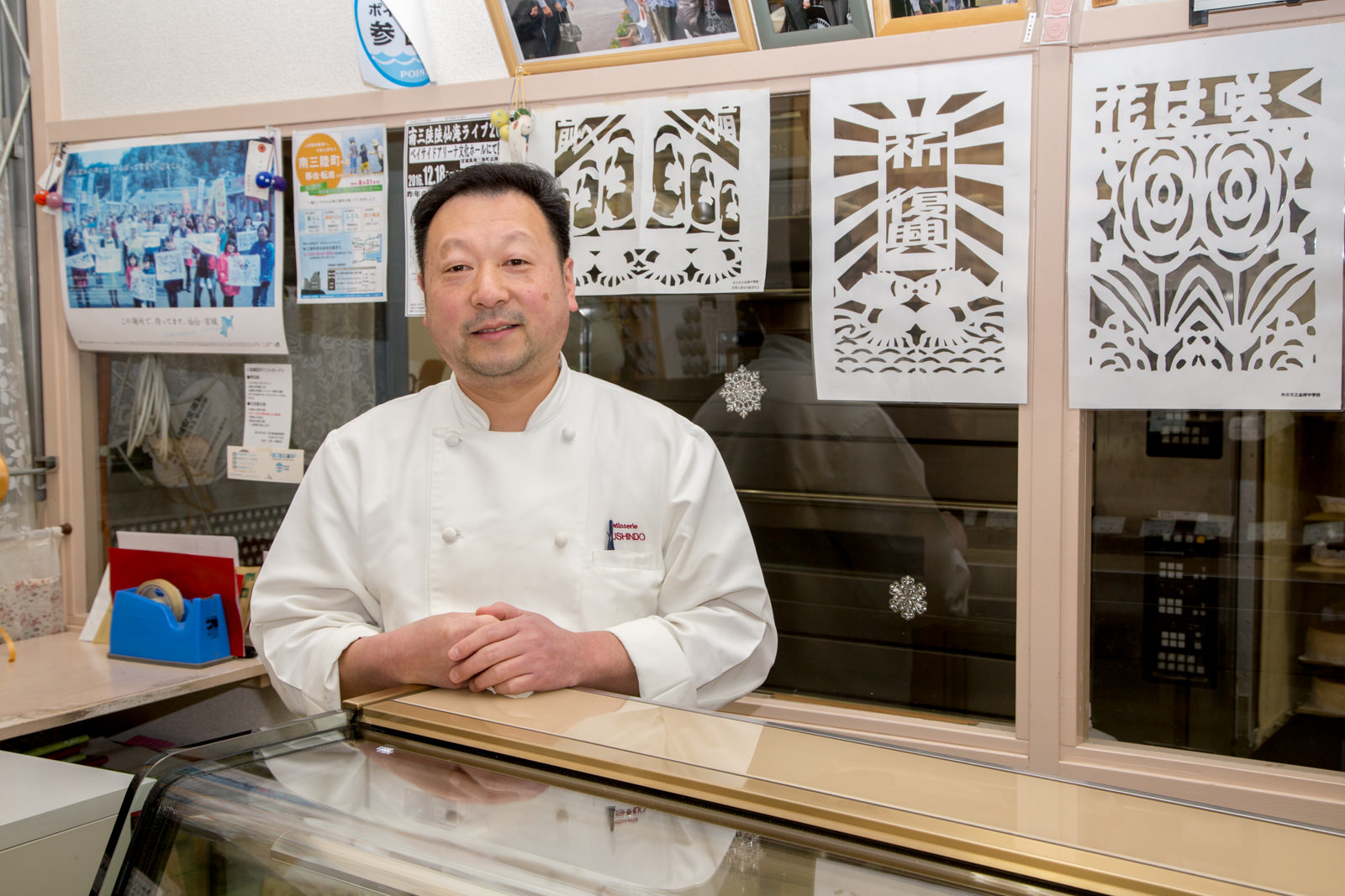 切り絵やポスターが貼られたガラス窓を背後に、ショーケース後ろに立っている、白いコック服を着た阿部雄一さんの写真