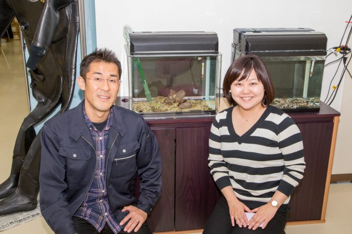 二つ並んだ水槽の前に座って笑顔でカメラを見ている阿部拓三さんと女性リポーターの写真