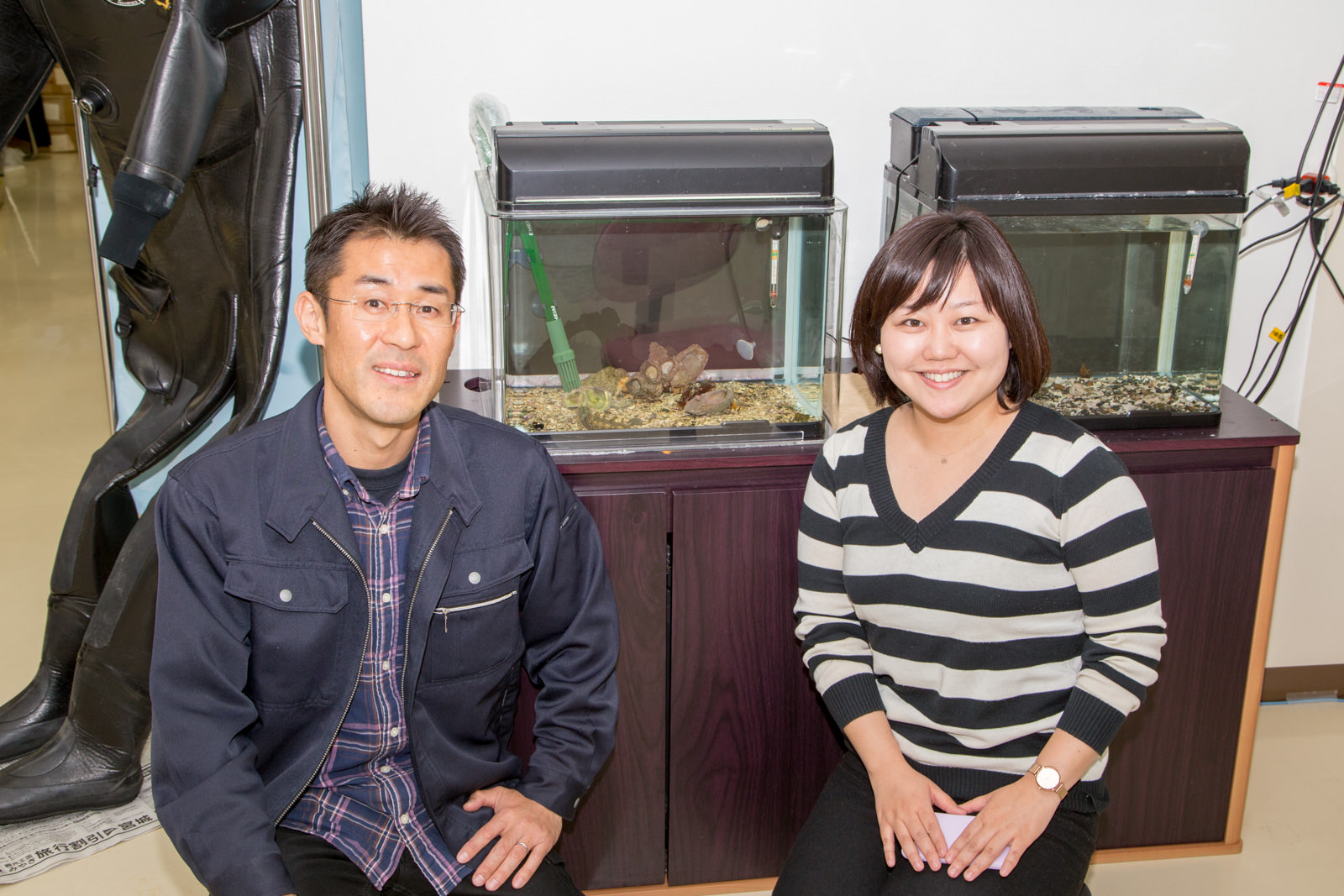 二つ並んだ水槽の前に座って笑顔でカメラを見ている阿部拓三さんと女性リポーターの写真