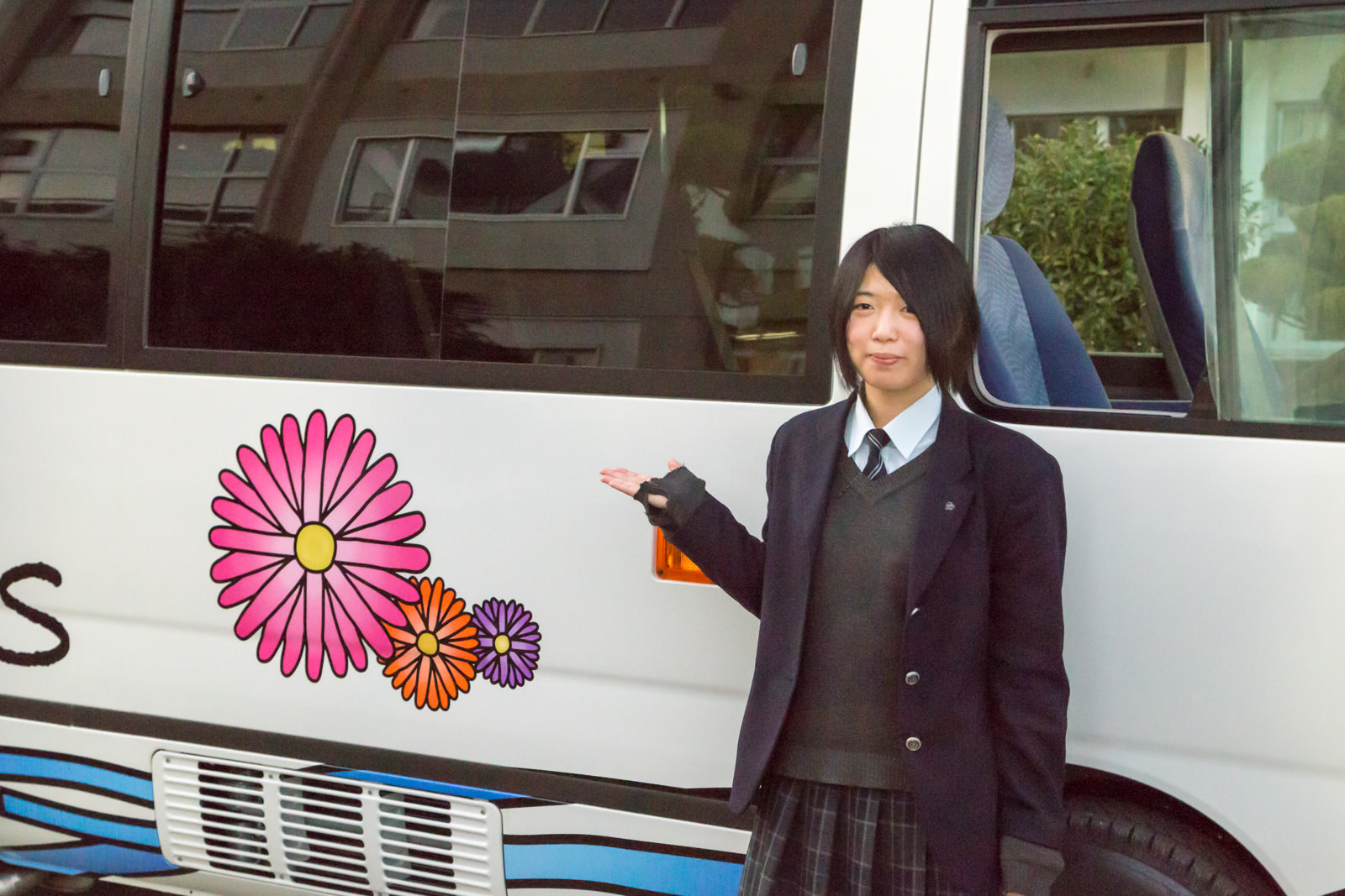 白地にカラフルな花のイラストが描かれたモアイバスの横で、制服姿の女子学生が嬉しそうに片手を差し出している写真