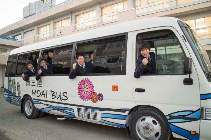 側面に「MOAI BUS」と書かれている白い小型バスにのった複数の男子高校生が、笑顔で窓から顔を出して元気にポーズを決めている写真