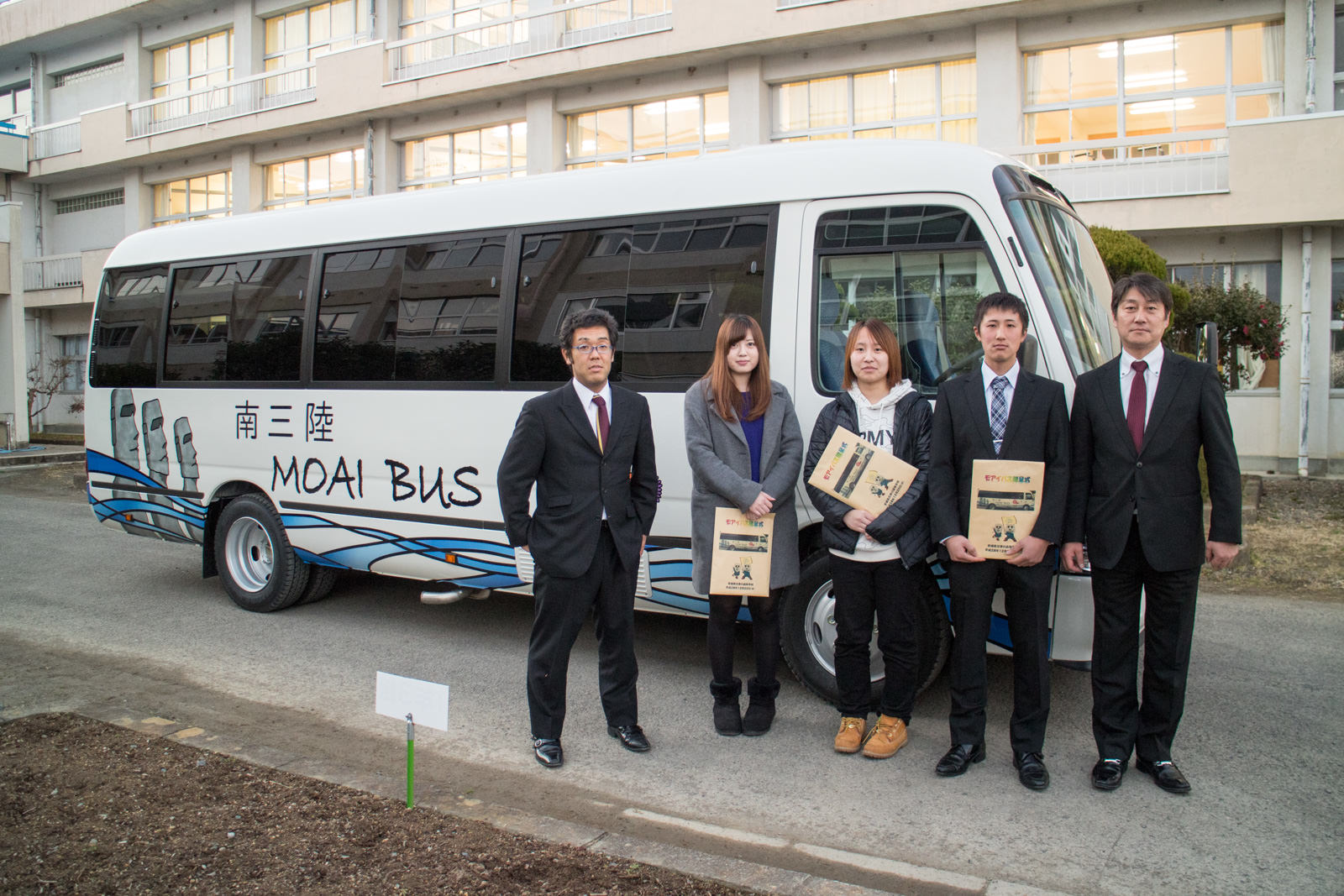 「南三陸 MOAI BUS」と書かれたバスの横に、スーツ姿の男性とそれぞれパンフレットを手に持っているカジュアルな服装の男女5人が並んでいる写真