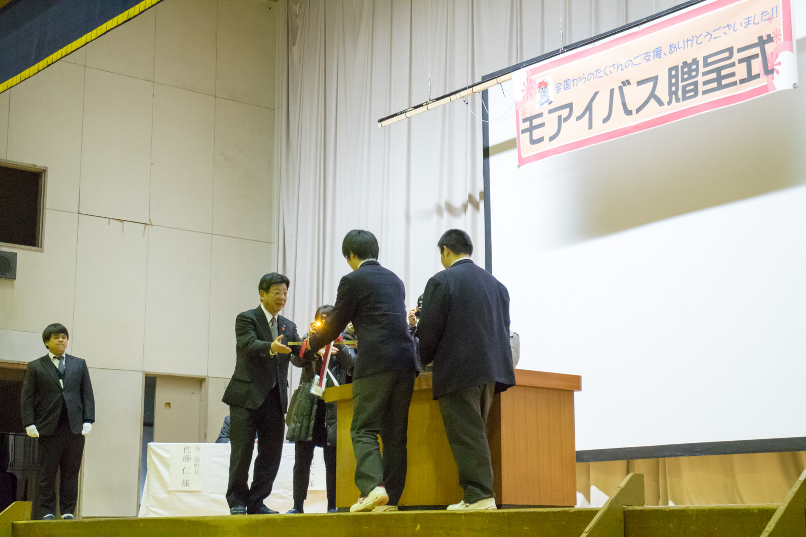 周囲には他の人々やカメラマンが見守っている式典の壇上で、スーツ姿の佐藤町長が制服姿の男子高校生からレプリカ鍵を受け取っている様子の写真