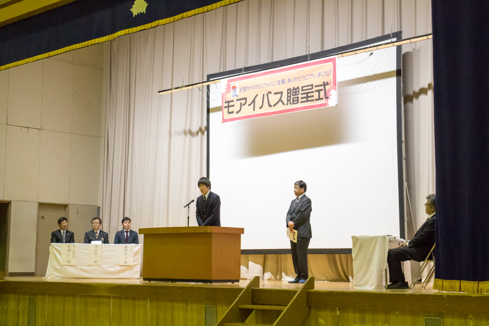 「モアイバス贈呈式」と書かれた横断幕が掲げられているステージの両側に数名の来賓者が座っており、壇上に立つ男子高校生がマイクの前で挨拶を行っている写真