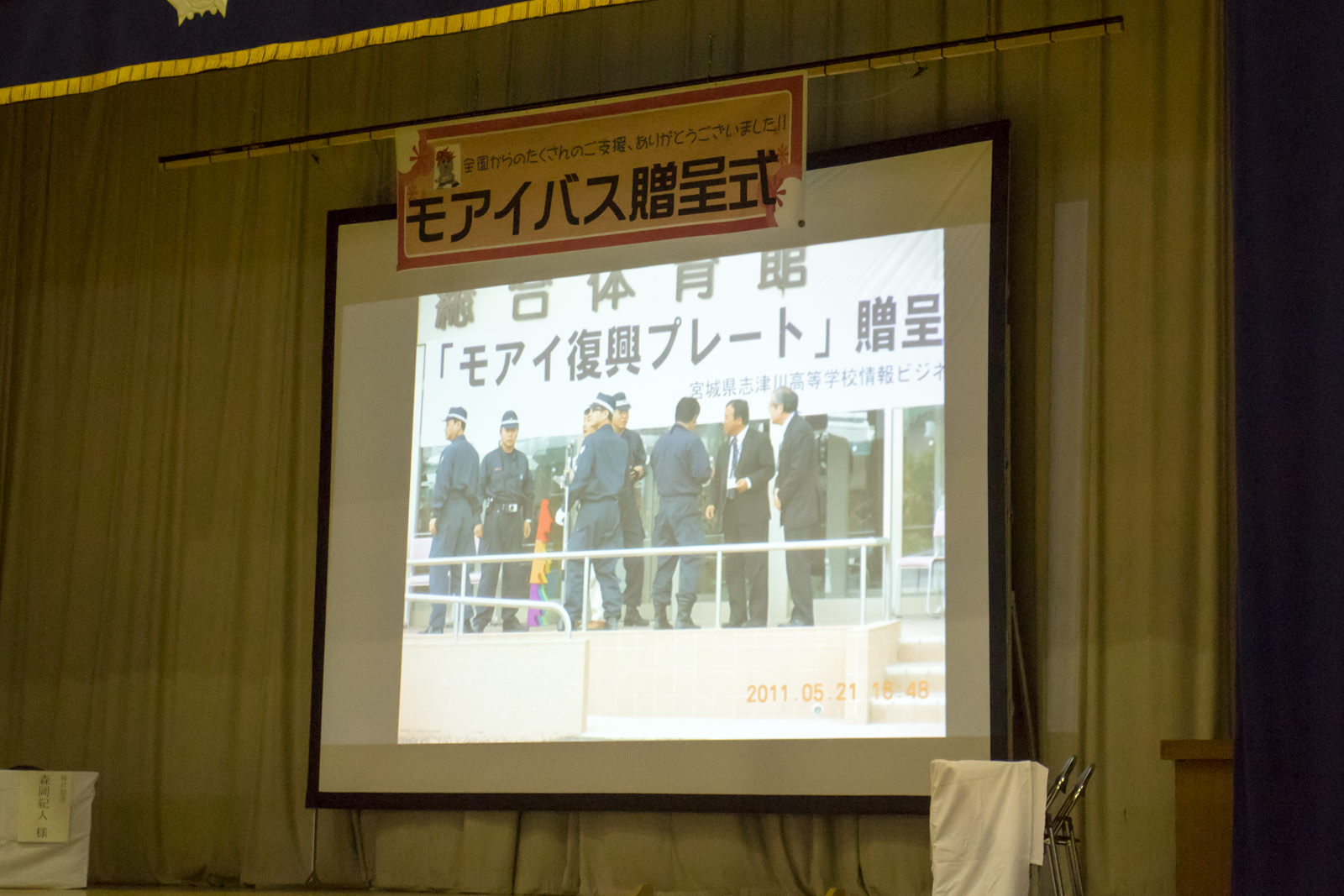 数名の警察官とスーツ姿の大人が建物の前に集まっており、「モアイ復興プレート贈呈」と書かれた贈呈の様子が映し出されたスクリーンをアップで撮影した写真