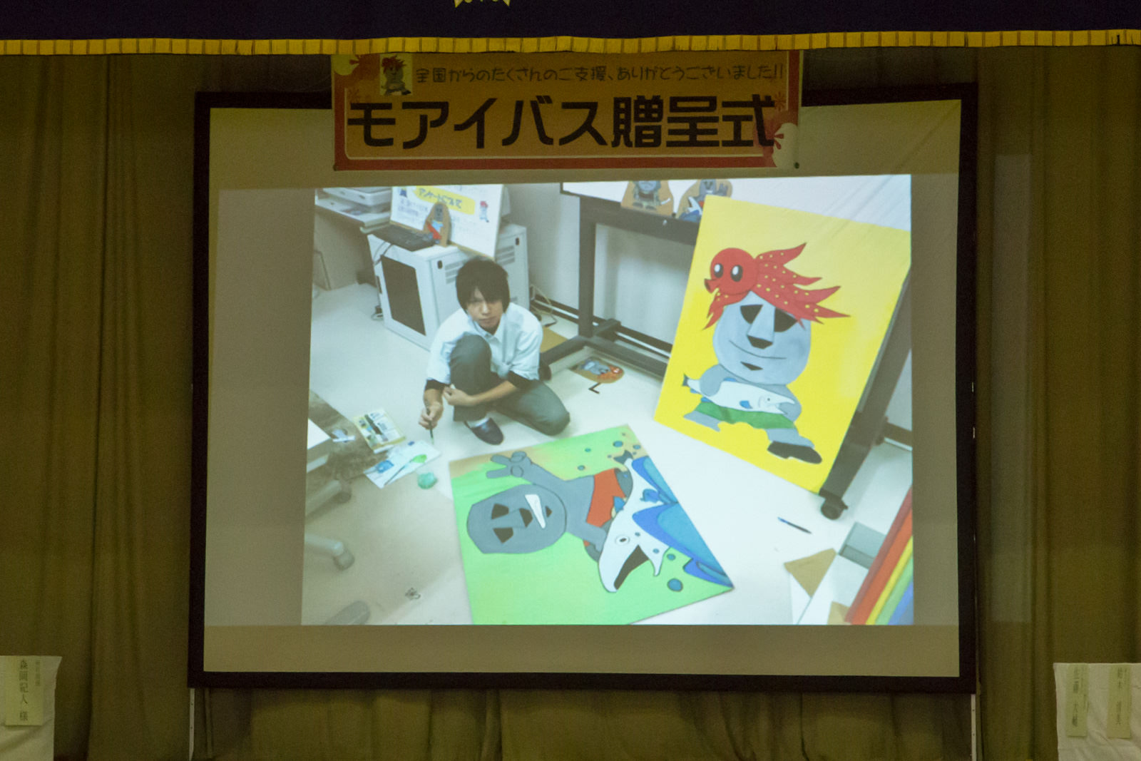 周囲にはカラフルなモアイ像をモチーフにしたイラストが描かれたパネルが数枚立てかけられている室内で、男子生徒が床に座って絵を描いている様子の写真