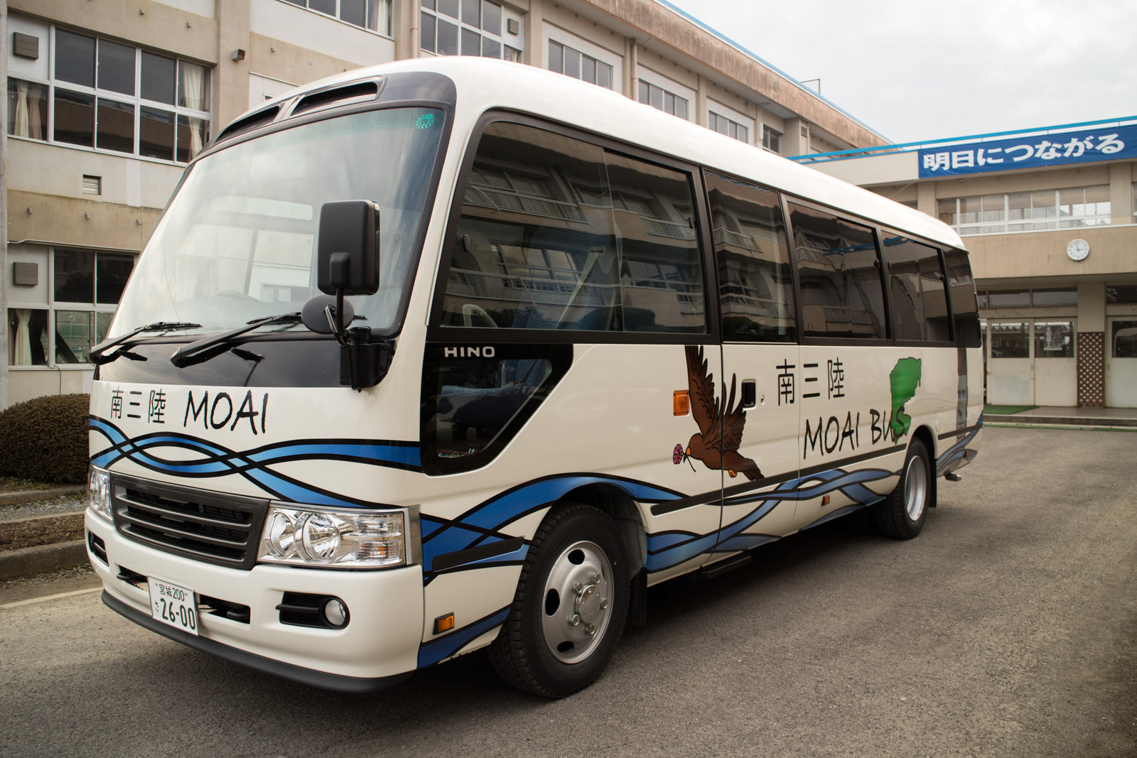 バスの側面にはワシと緑の地図のイラストが描かれており、白地に「南三陸 MOAI BUS」と書かれたモアイバスが校舎の前に停まっている写真
