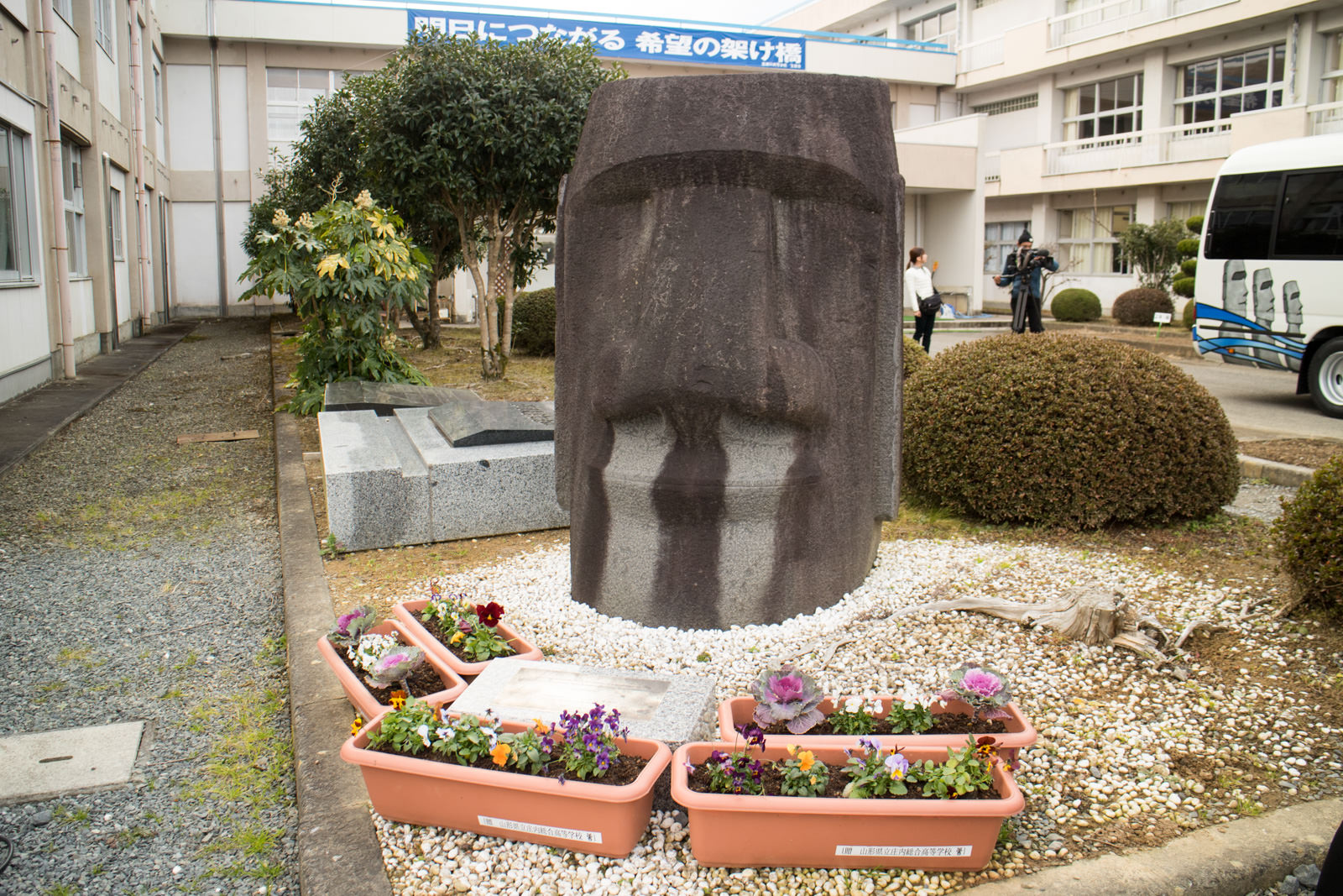 校舎に囲まれた中庭に大きなモアイ像が設置されており、像の周囲には花が植えられたプランターや白い小石が並べられている写真