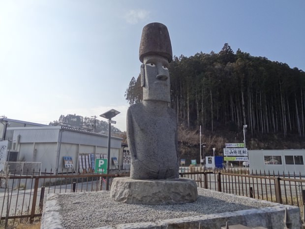 木々に囲まれ、仮設建物が建っている前に、目が入っている灰色のモアイ像が立っている写真