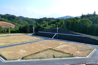 舗装された道路と電柱が整備された新しい住宅地に、まだ建物が建っていない区画が並び、背景には森林の丘と遠くの山々が広がっている造成地の写真