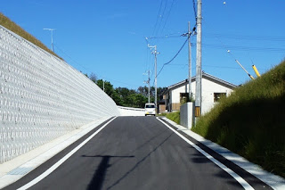 左に擁壁、右に電柱と住宅が並ぶ見通しの良い坂道が続く新興住宅地の道路の写真