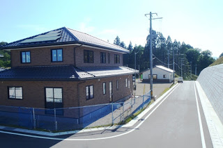 茶色の外壁の二階建ての住宅のそばに舗装された道が通り、周囲に開発中の区画が見える住宅地の写真