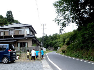 道路横の駐車スペースに数名の人たちが立っており、その奥に2階建ての家が建っている写真