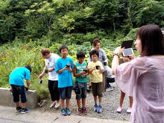 2名の女性と4名の男の子が屋外の花壇の前で黒い苗ポットを手に持って立っており、スマートフォンを顔の前で持った水華里さんがその様子を撮影しようとしている様子の写真