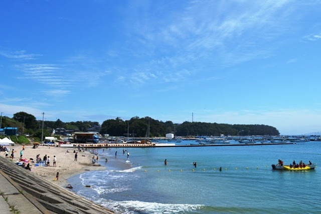 防波堤沿いに広がる白い砂浜と青い海に、多くの人々が水遊びやボートを楽しんでいる夏の海辺の風景が写っている写真
