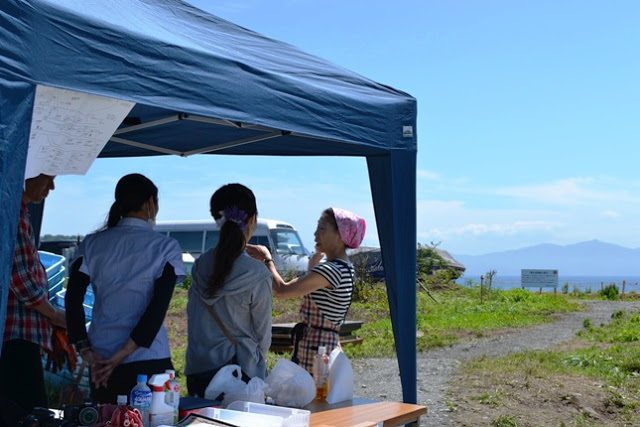 海が後ろに見える場所に建てられた青いテントの下で女性たちが集まり、ピンクの三角巾を頭につけ、エプソン姿の女性の話を聞いている様子の写真