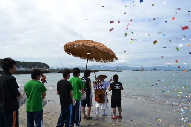 砂浜で緑や黒のTシャツを着た人々が、伝統的な衣装を着た人物とともに海を見ながら紙吹雪が舞う中で儀式のような行事に参加している写真
