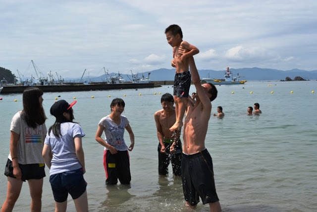 海の中で男の子を高く持ち上げて遊んでいる男性と、それを笑顔で見守る若者たちが写っており、楽しそうな雰囲気が伝わる写真