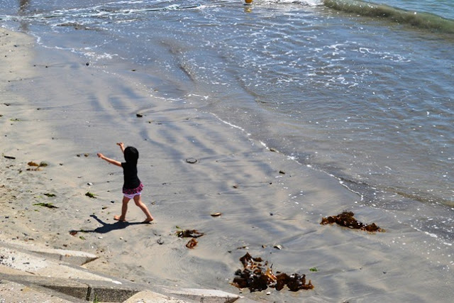 砂浜でひとりの幼い女の子が手を広げながら歩いており、すぐそばに静かに波が打ち寄せている様子を写した写真