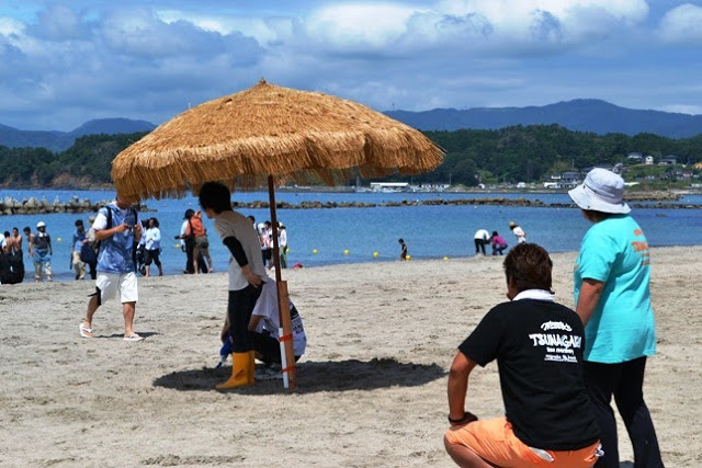 ビーチ中央にある藁で作られた大きな日除けの下で2人の男性が座って海を見ている様子を写した写真